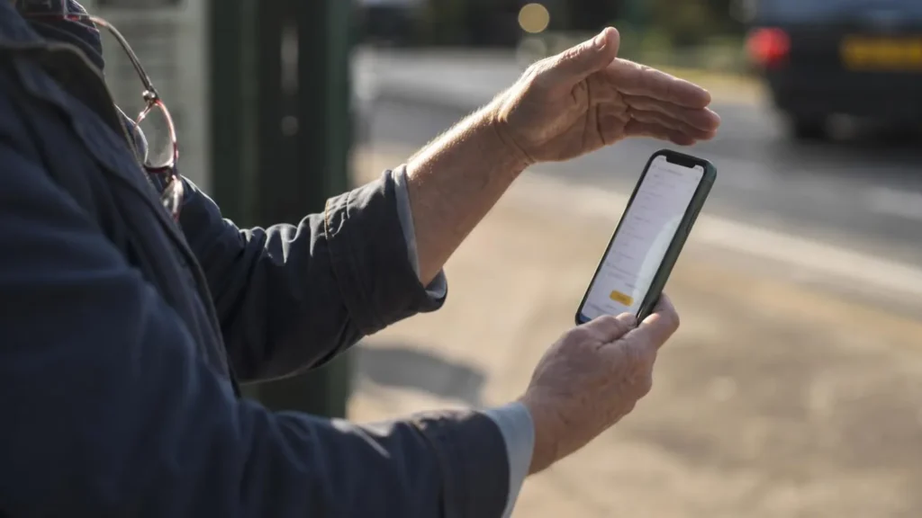 An older person at a roadside holding a phone at arm's length in bright sun, shading the washed-out screen with their other hand, reading glasses hooked on their collar.